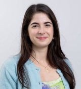 Eleonora, a white young woman with dark brown hair and eyes, smiling and wearing a green/purple top and a light blue sweater.