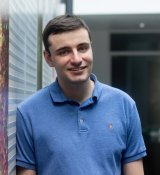 Portrait photo of Adam Pavlinek leaning against a wall. He has dark brown hair, is smiling, and wearing a blue polo shirt.